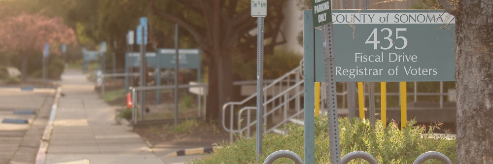 Picture of the sidewalk outside of the Registrar of Voters Office at sunset. A large green sign at the right says, "County of Sonoma 435 Fiscal Drive Registrar of Voters."